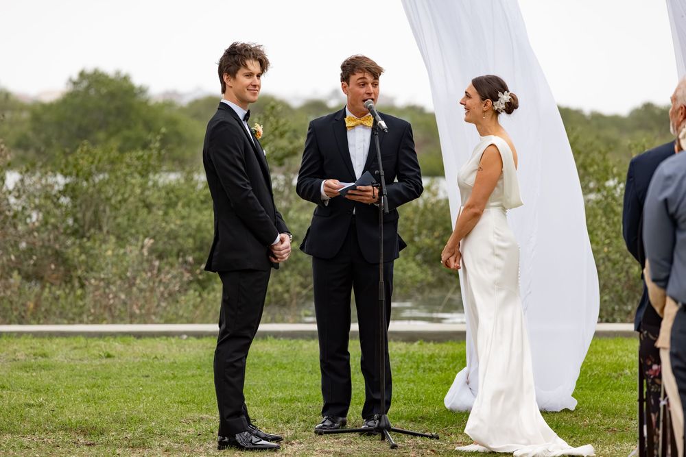 Outdoor wedding ceremony with groom, officiant reading vows, and bride in white gown standing by decorative white drape.