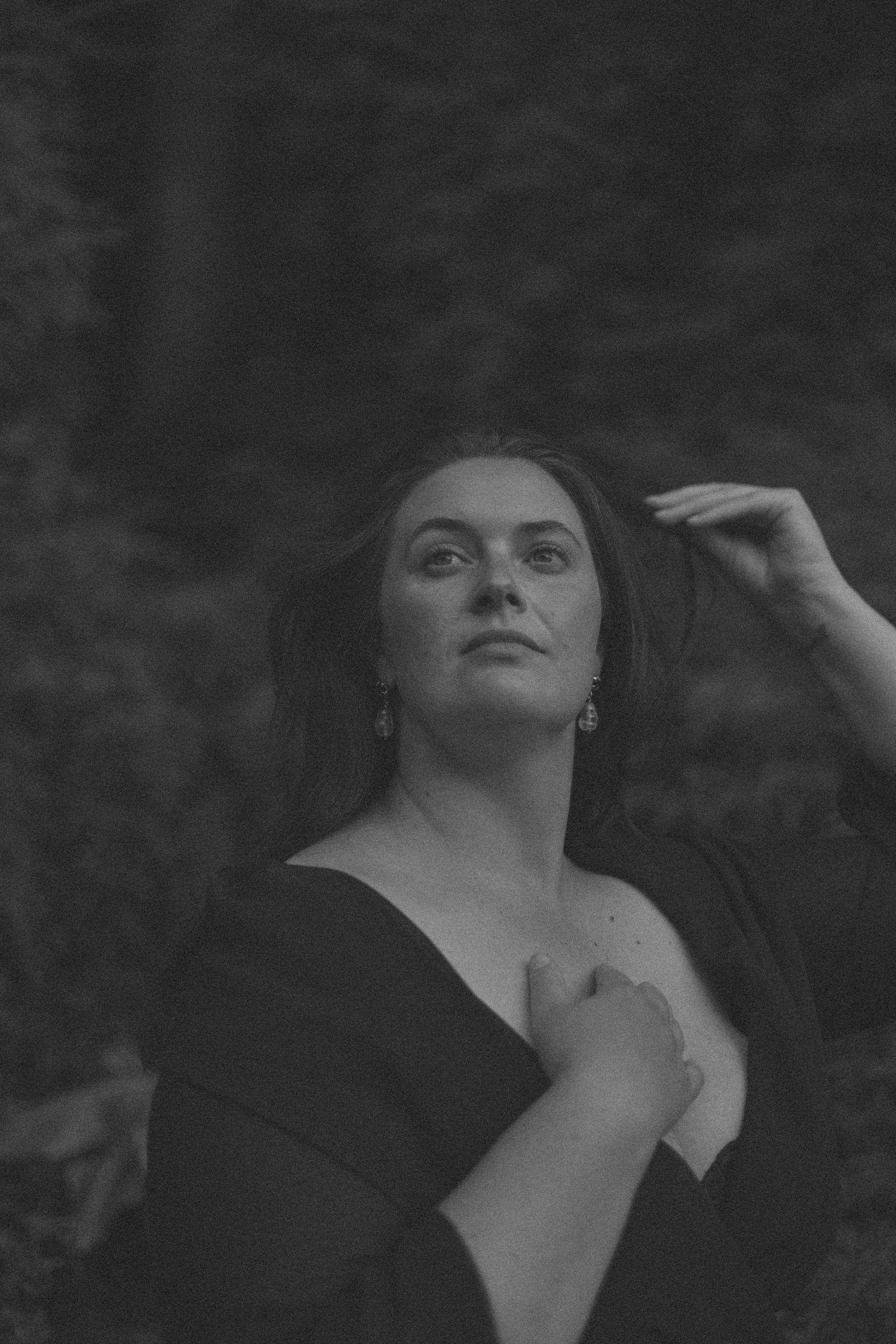 Black and white portrait of a woman in a black dress touching her hair with a dreamy forest backdrop in New Zealand.