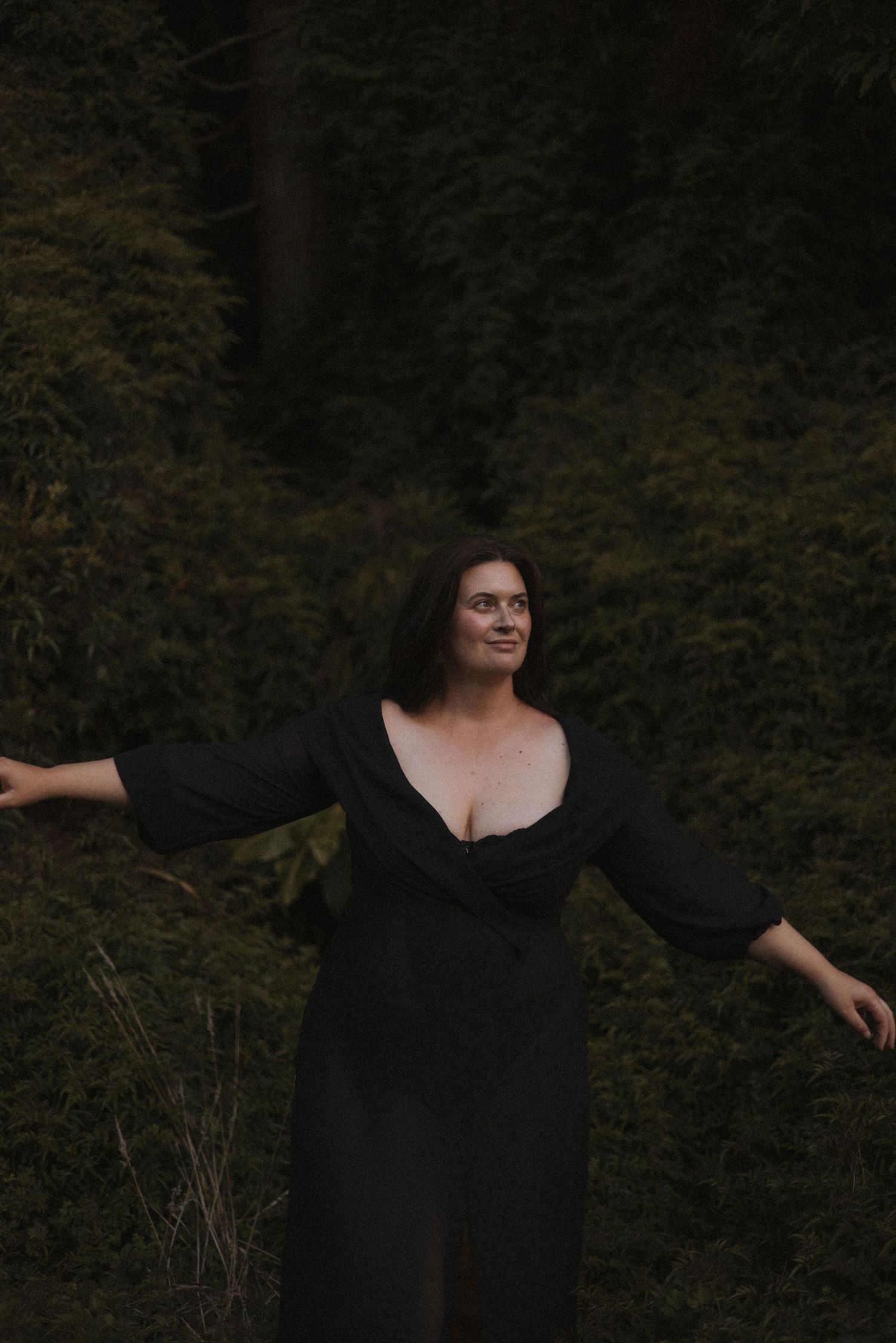 Woman in black wrap dress standing with arms outstretched smiling in front of lush green trees in New Zealand.