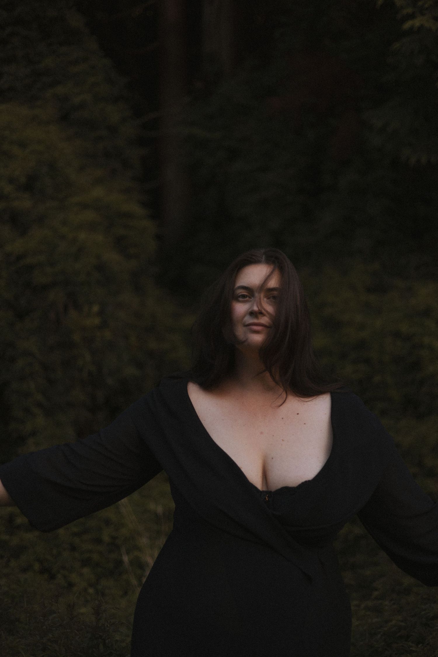 Woman in black deep-V wrap dress posing confidently in front of dark green forest foliage in New Zealand.