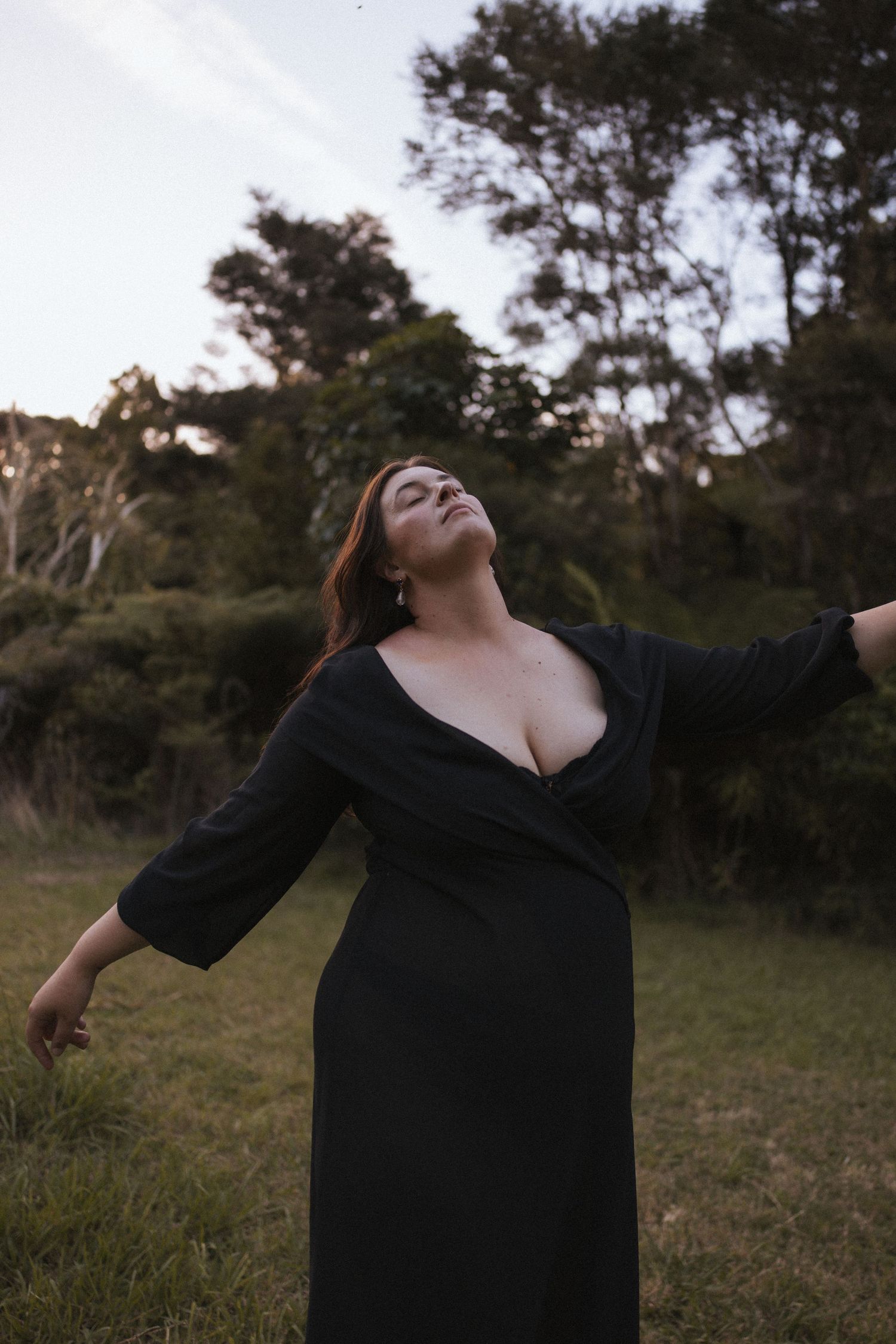 Woman in black wrap dress with arms spread wide and eyes closed in an open grassy field at dusk in New Zealand.