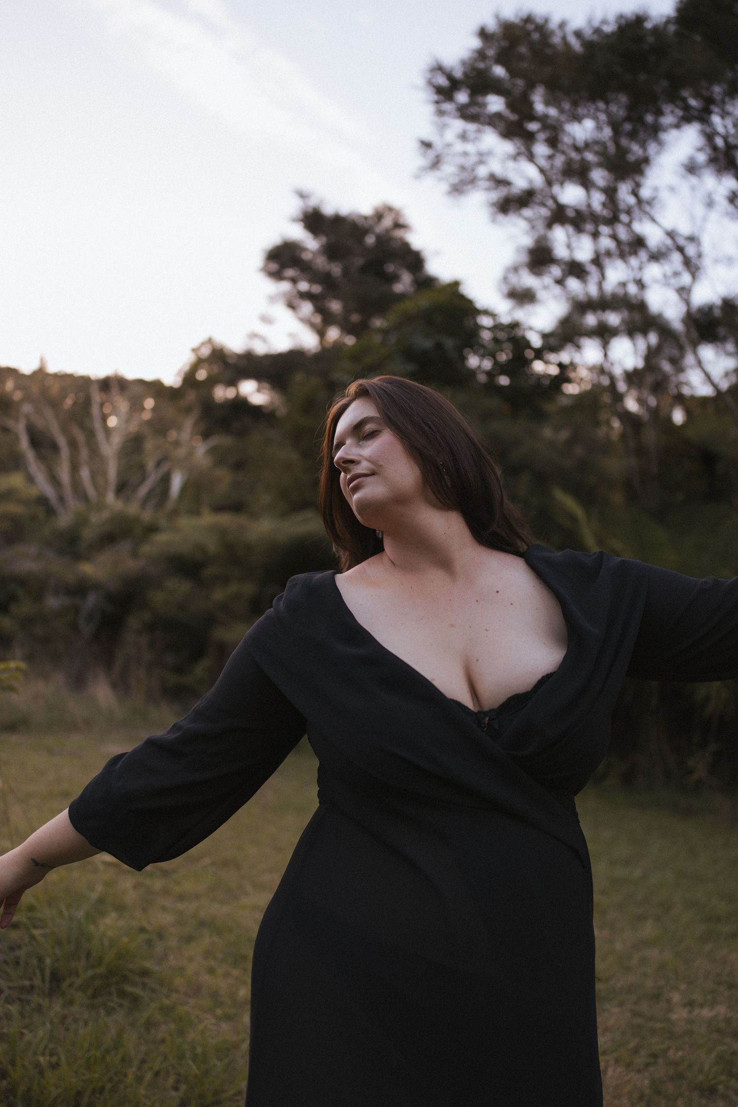 Woman in black wrap dress with arms outstretched and eyes closed in a peaceful outdoor grassy setting in New Zealand.