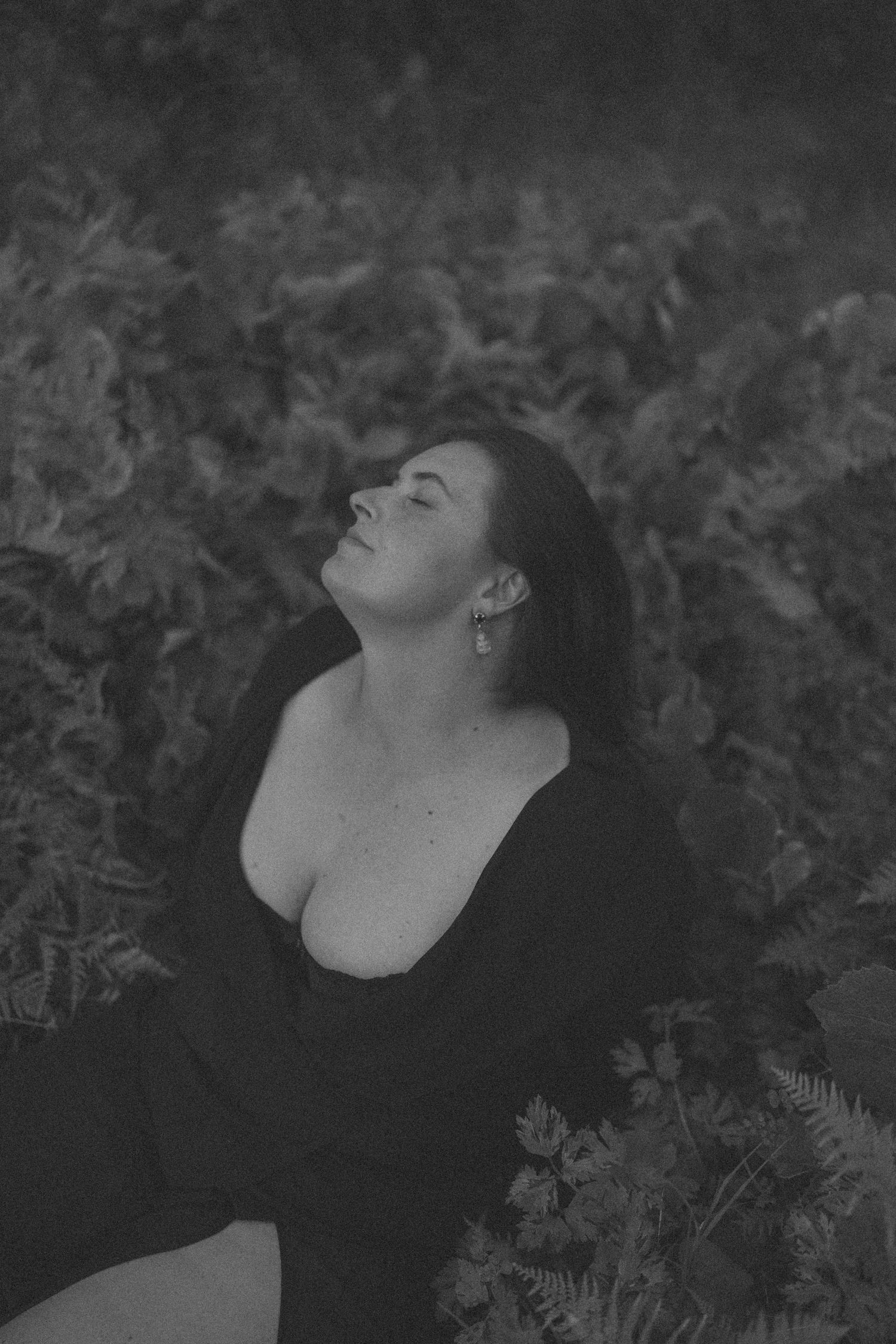 Black and white portrait of a dark-haired woman in black dress sitting among ferns, head tilted back in New Zealand.