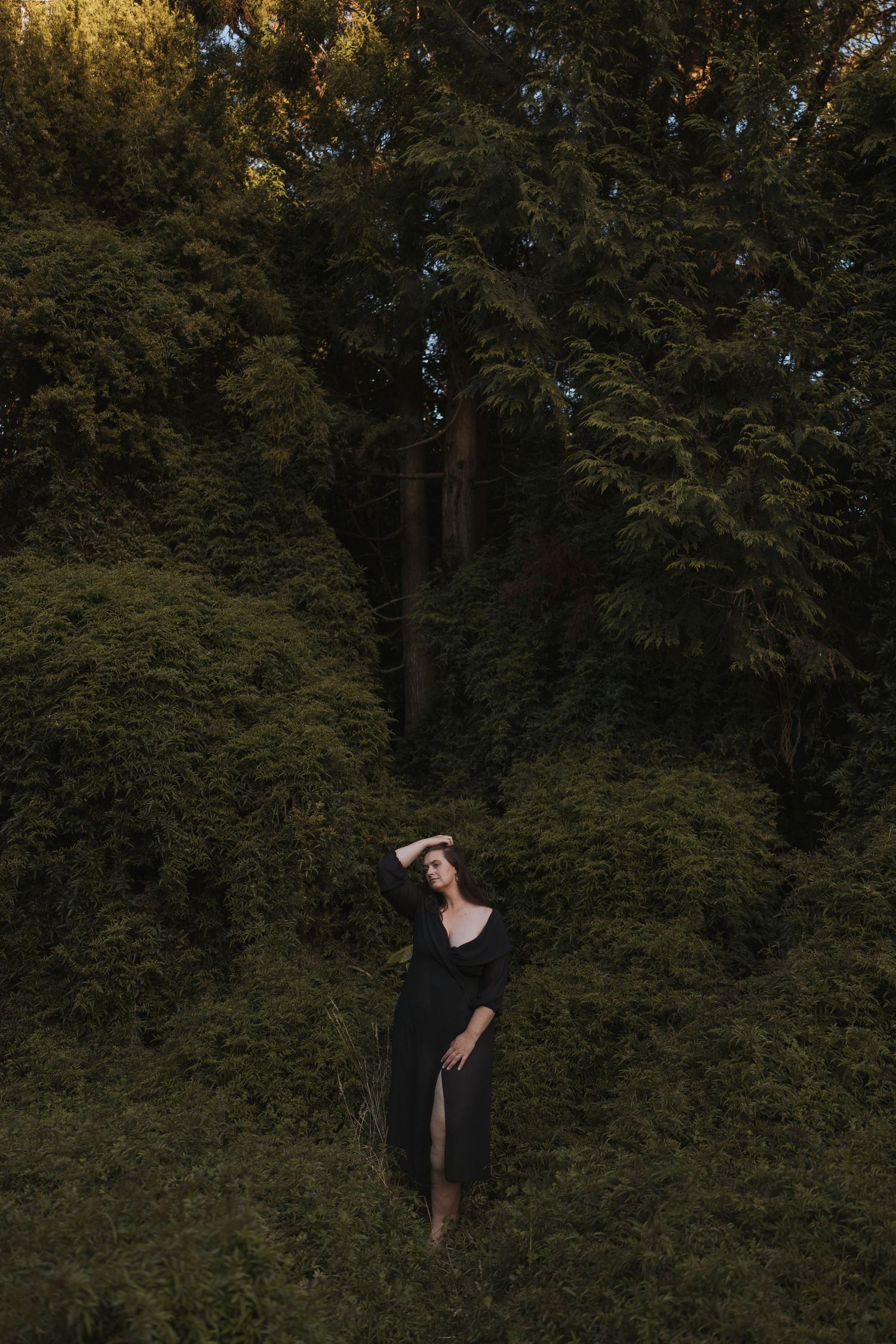 Woman in elegant black dress posing confidently in front of dense green trees at dusk outdoors in New Zealand.