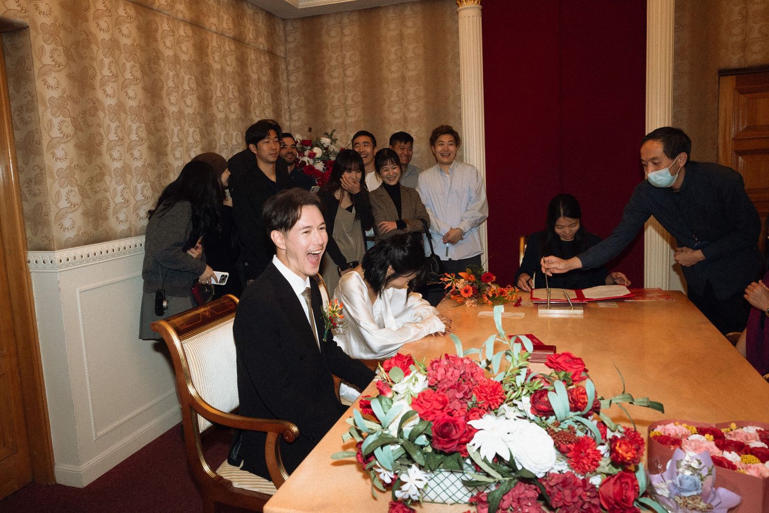 Couple signing marriage documents at a registry office ceremony surrounded by guests and red floral decorations.