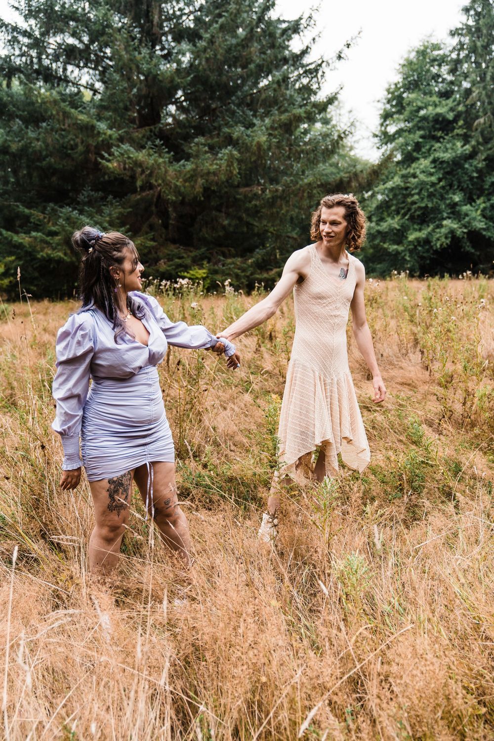 Two women in dresses hold hands walking through a golden field with evergreen trees in the background. olympic national park elopement