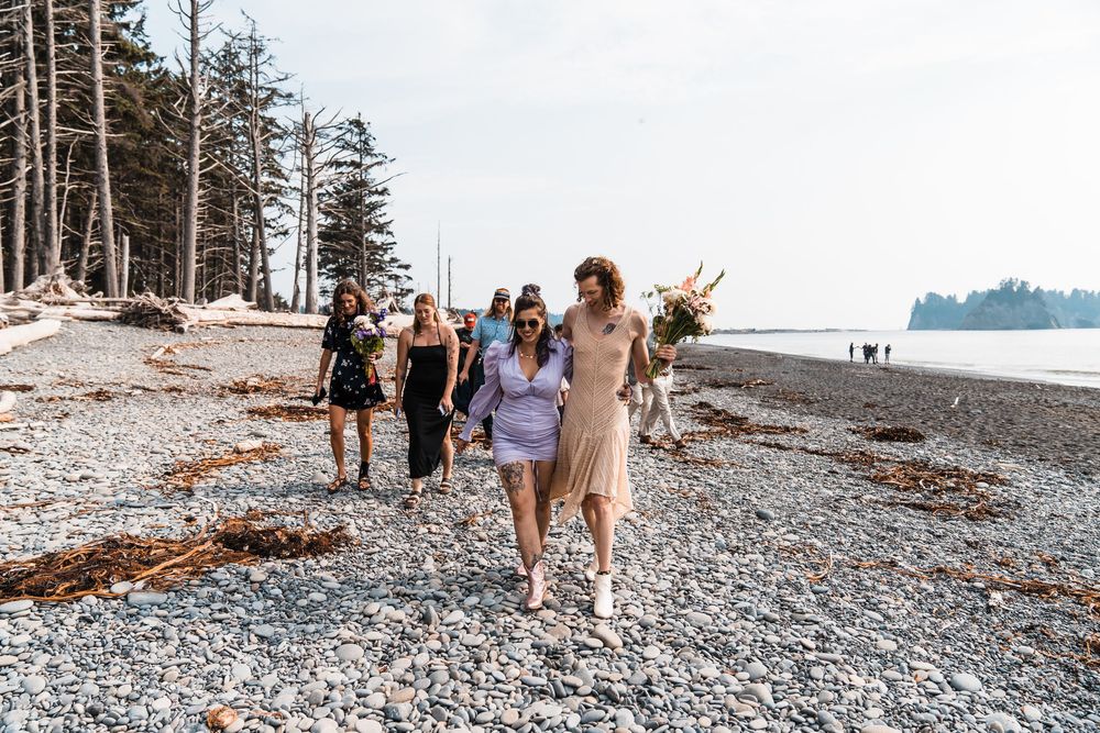 Group of women in summer dresses walking on a rocky beach surrounded by tall pine trees on a sunny day. olympic national park elopement