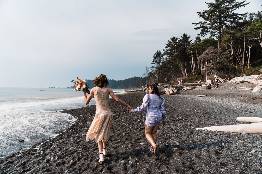 Two women run hand-in-hand along a rocky Pacific Northwest beach, one holding a bouquet, with driftwood and evergreens behind. olympic natio