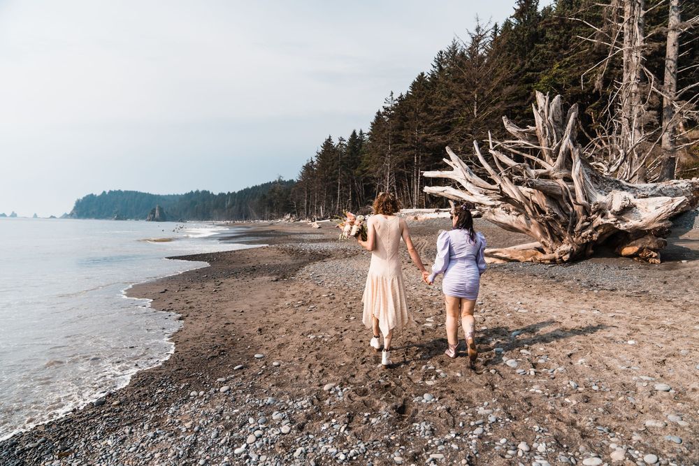 Two women walk along a rocky Pacific Northwest beach with driftwood and evergreen trees. olympic national park elopement