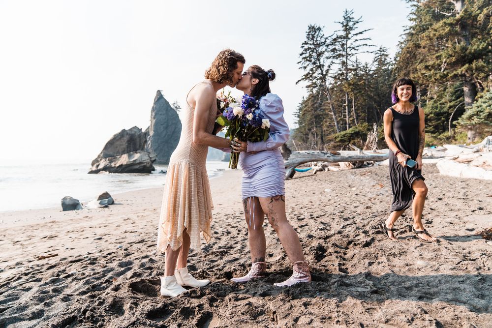Two brides share a kiss on a sandy beach during an outdoor wedding ceremony with a witness nearby. olympic national park elopement