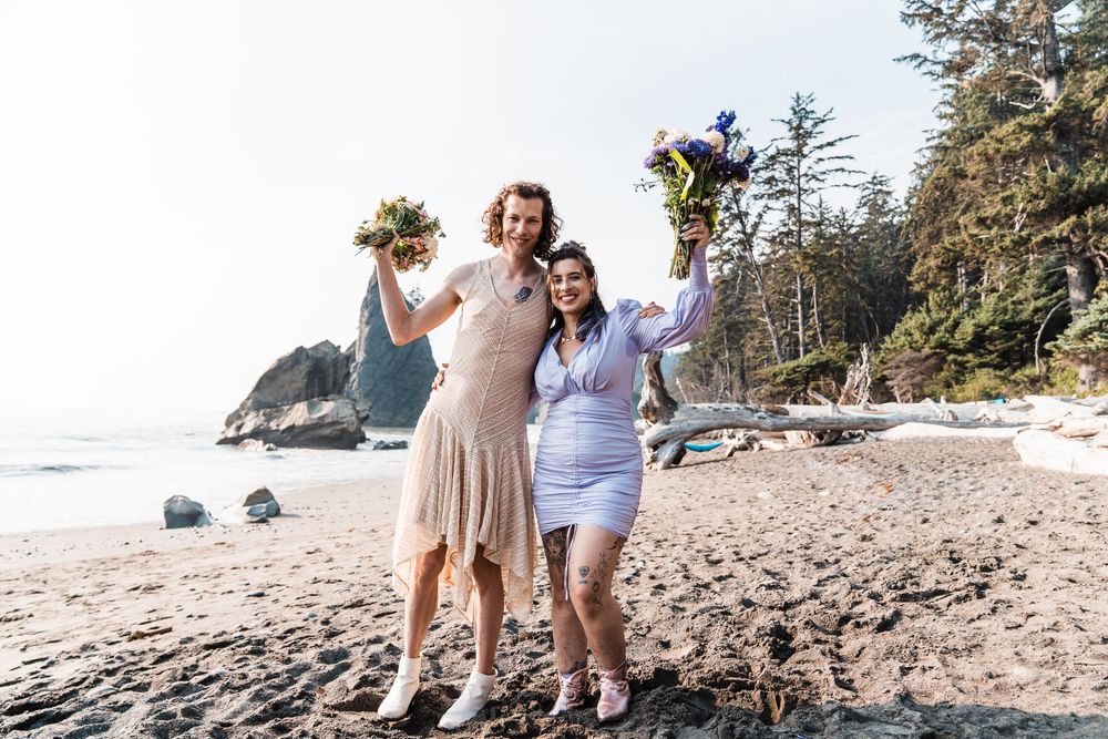 Two brides celebrate on a scenic Pacific Northwest beach, holding colorful bouquets aloft with sea stacks in the background. olympic nationa