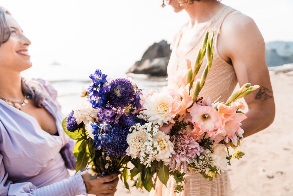 Two women holding colorful bridal bouquets of purple, blue, and pink flowers on a beach.olympic national park elopement