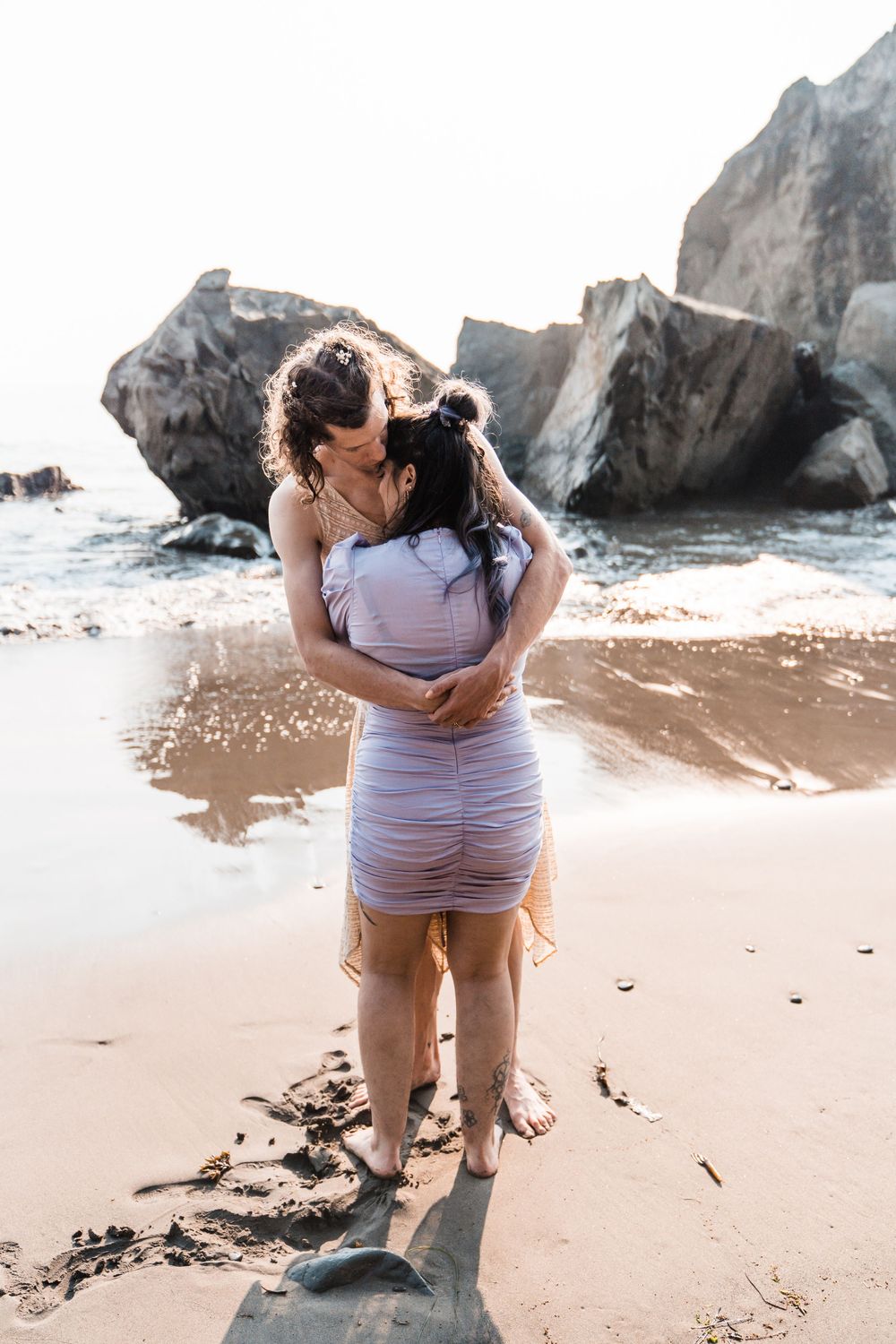 Two women embrace and kiss on a sandy beach with large rocks and ocean waves in the soft golden light. olympic national park elopement