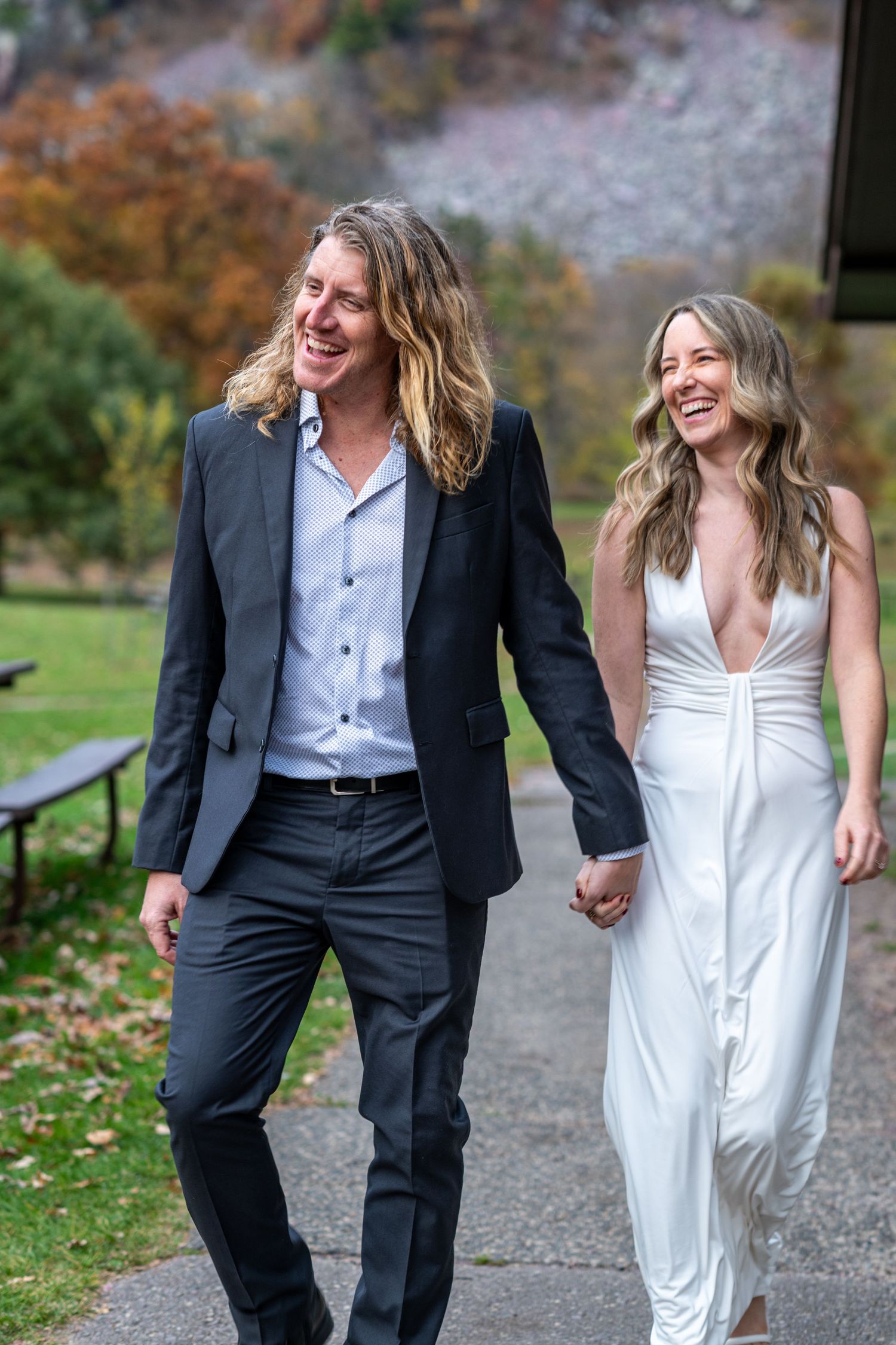 Happy couple walks hand-in-hand outdoors, bride in white gown and groom in dark suit, autumn trees in background.