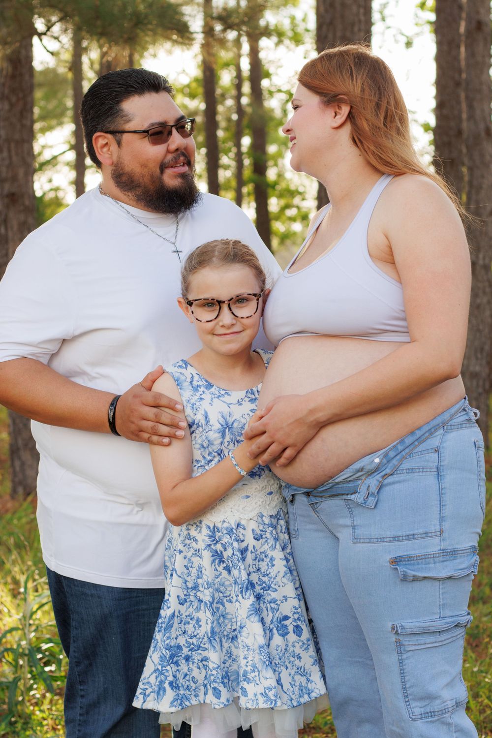 A pregnant woman, man, and young girl pose together outdoors in a forest for a family maternity photo.