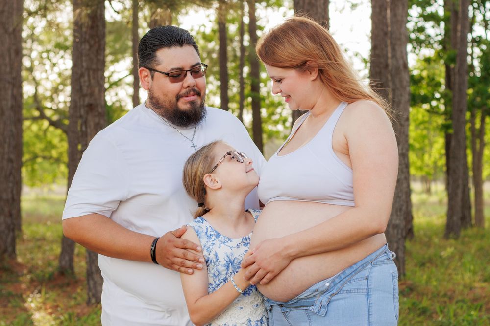 Pregnant woman and man embrace young girl with glasses outdoors in a pine forest during a family maternity photo session.