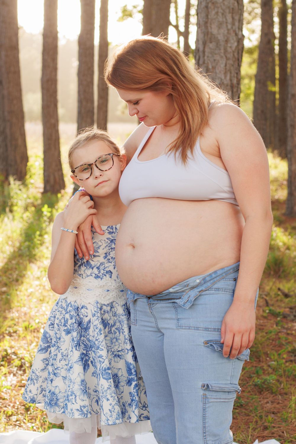 Pregnant woman in white crop top and jeans stands with young girl in floral dress in sunny forest setting.