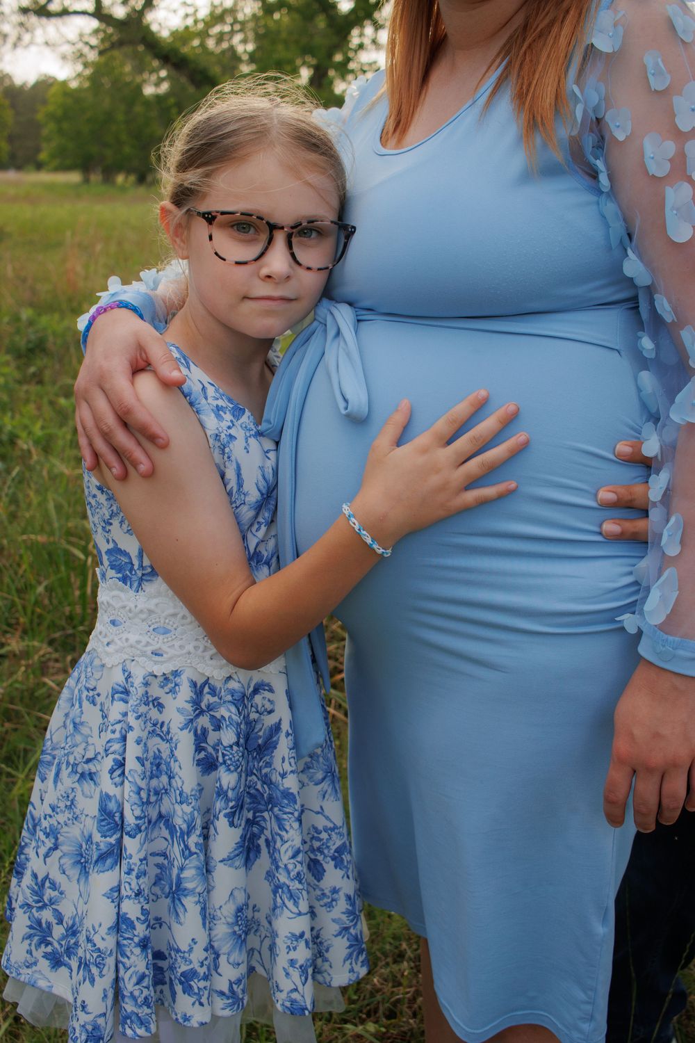 Young girl in glasses hugging pregnant woman in blue dress outdoors in a green field.