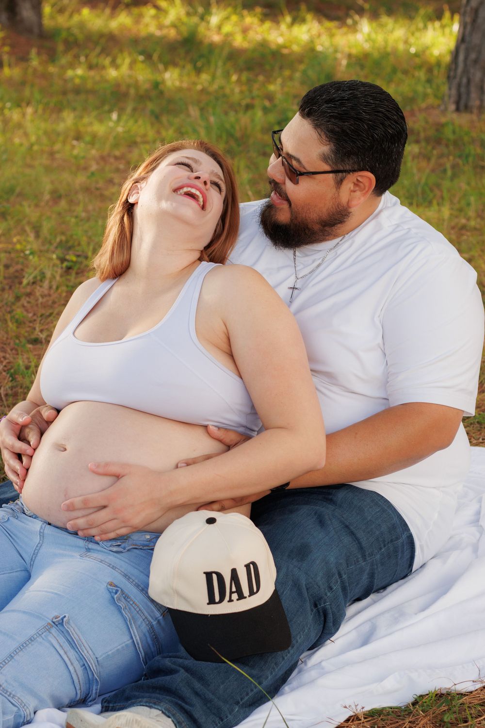 Pregnant woman smiles while sitting outdoors with her partner, who holds a DAD cap on her baby bump.
