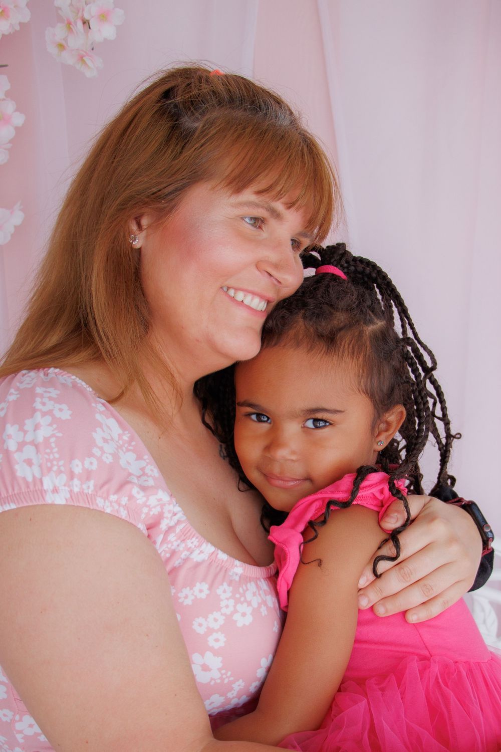 A smiling woman in pink floral top hugs a young girl with braids in a pink tutu against a pink backdrop.