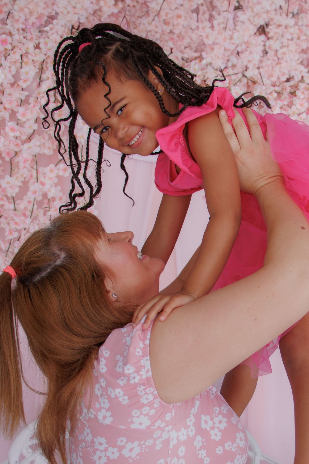 A woman lifts a smiling young girl in a pink dress against a floral pink background.