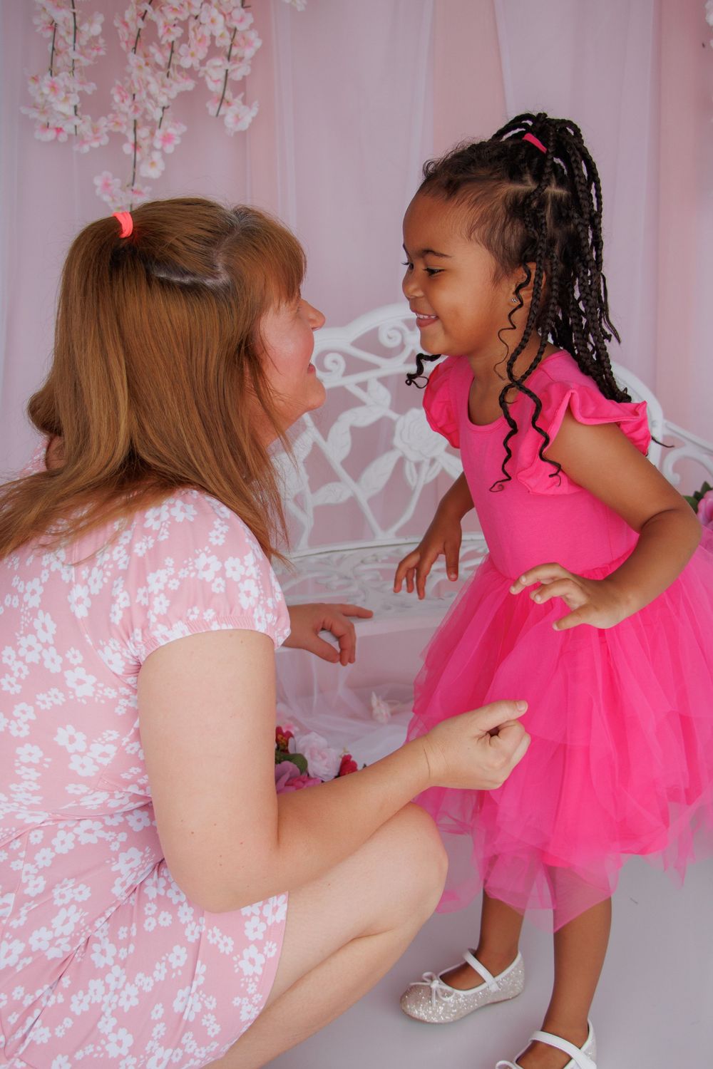 Woman in floral dress kneels to interact with smiling girl in pink tutu dress against pink backdrop.