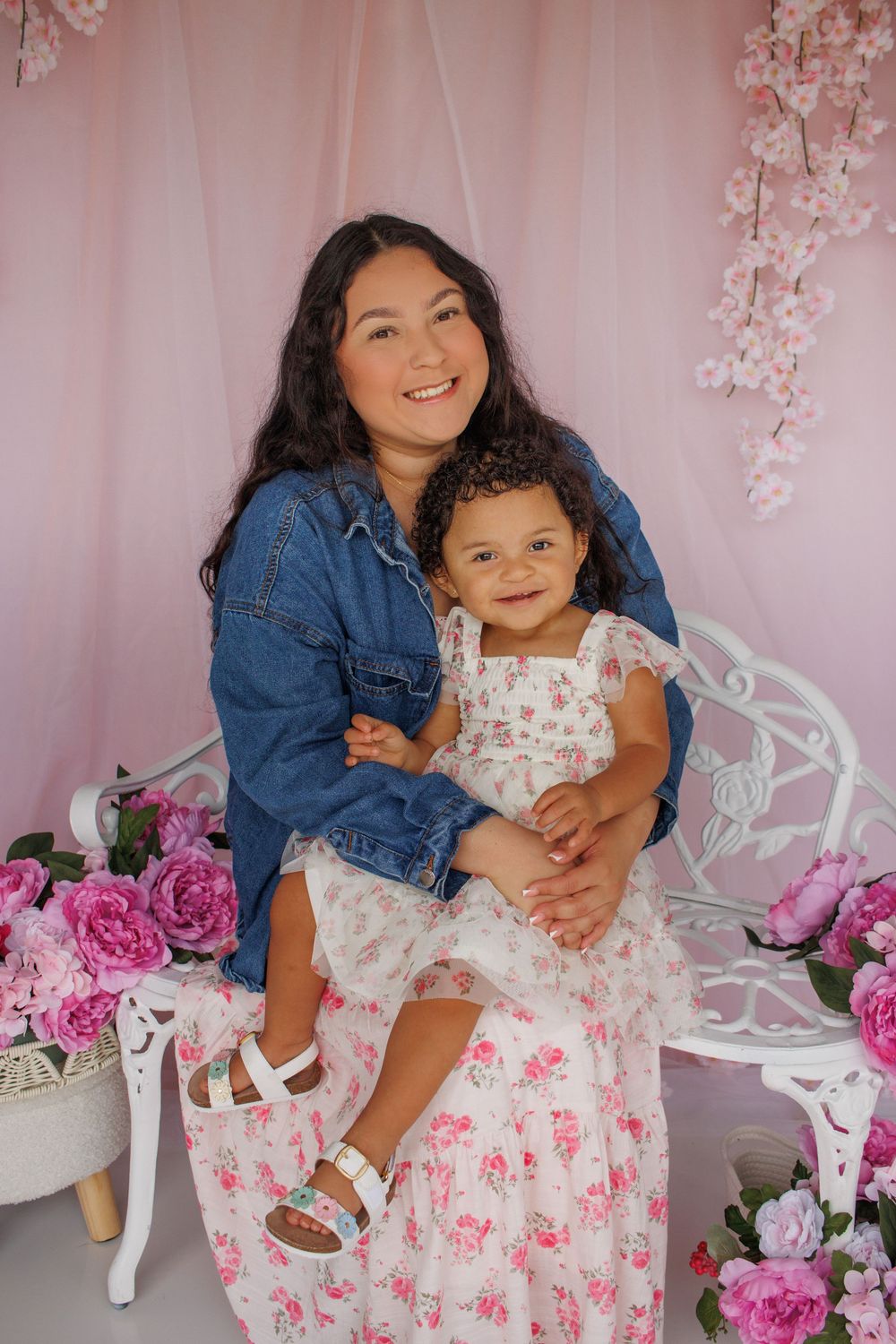 A woman in a denim jacket holds a toddler girl in a floral dress, posing on a white bench with pink flowers.