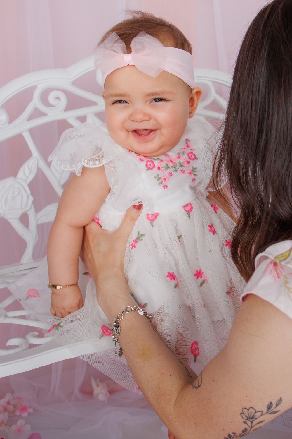 Smiling baby girl in white floral dress and pink bow headband being held by mother in pink studio setting.