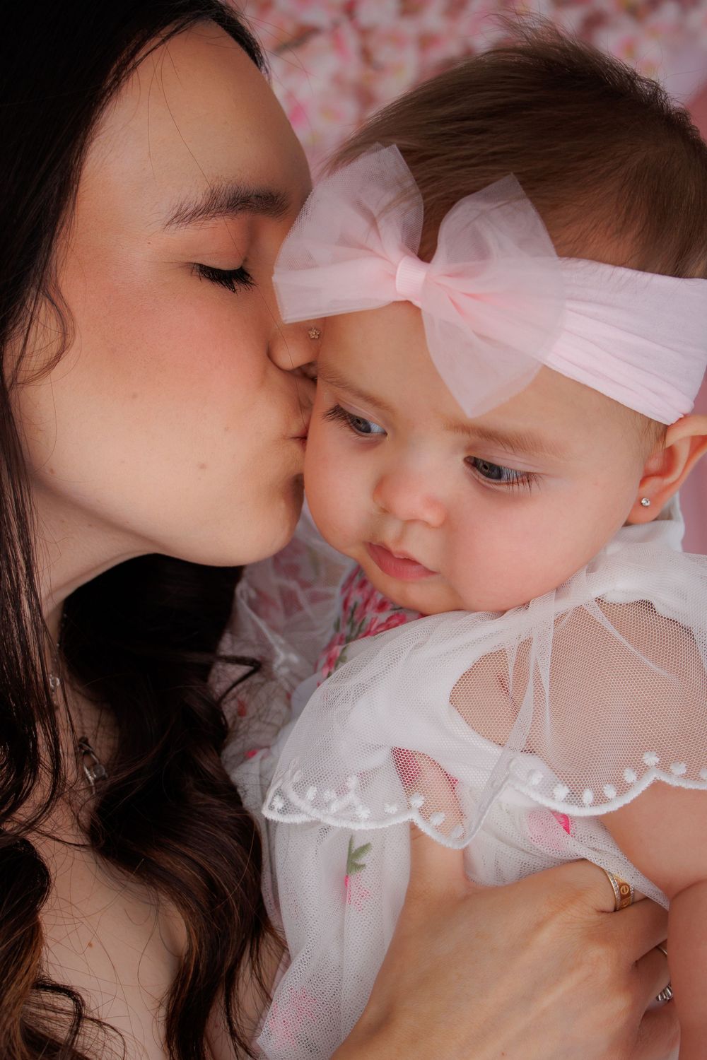 A loving mother kisses her baby girl wearing a white dress and pink bow headband in a sweet portrait.