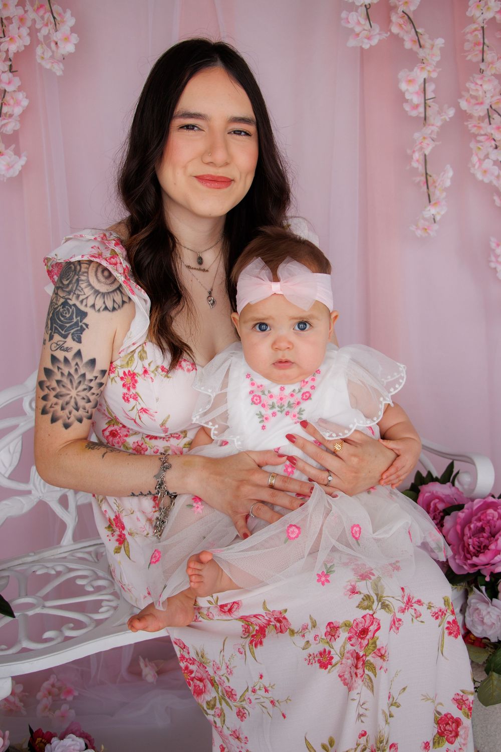 Young tattooed mother in floral dress holds baby girl in white dress with pink bow on white bench.