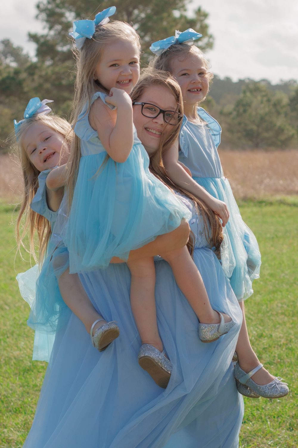 The image shows three young girls wearing matching blue dresses and butterfly hair accessories, smiling and posing together in a field.