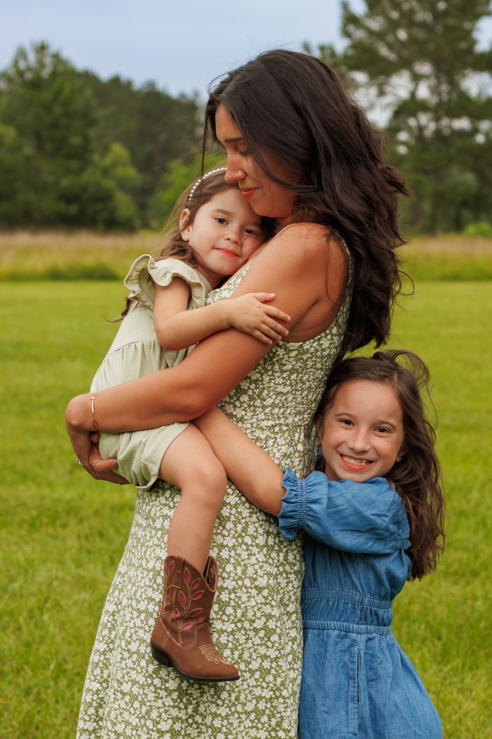 A mother in a floral dress holds her toddler while her older daughter hugs her in a green field.