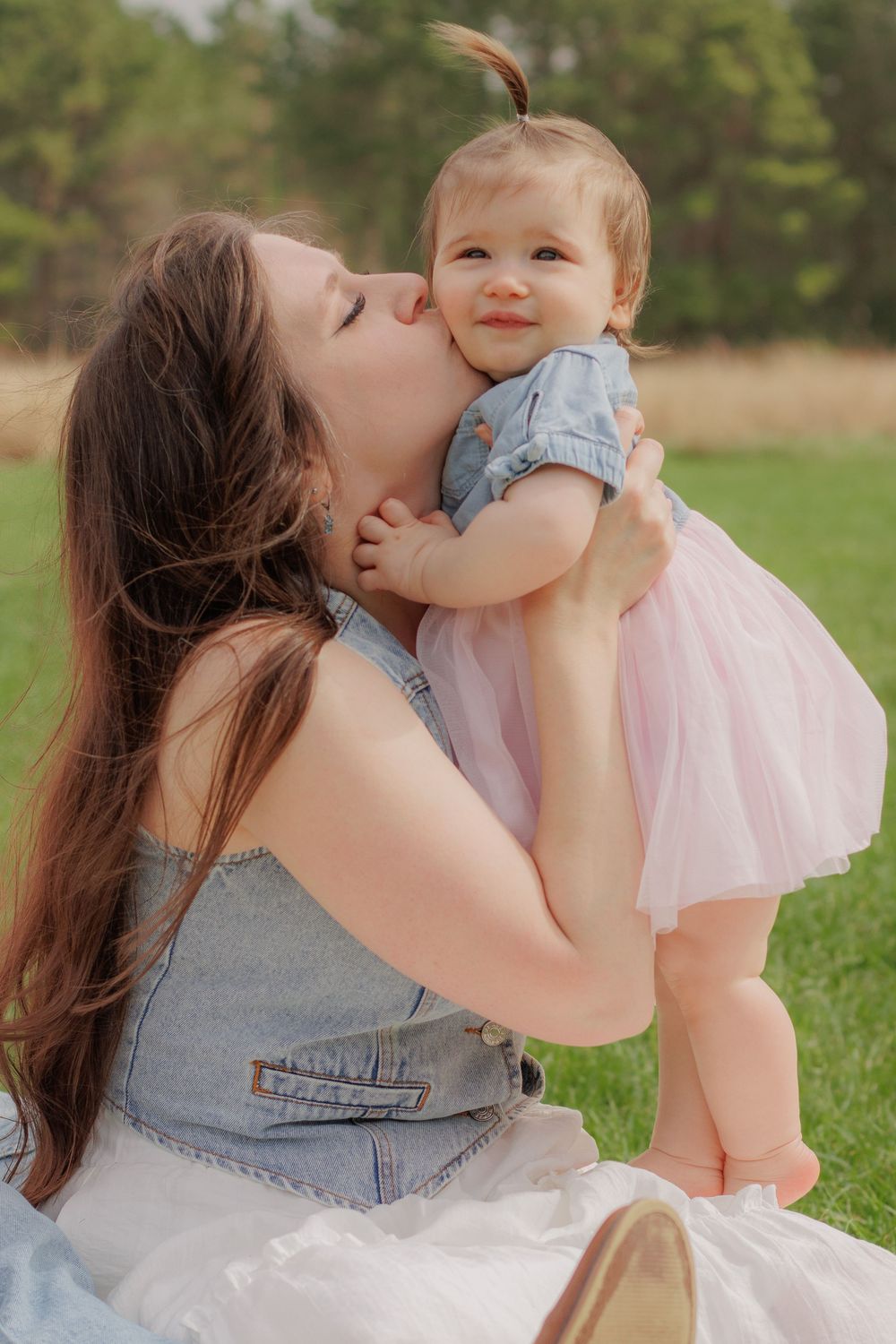 A loving mother kisses her smiling baby girl in a pink tutu skirt outdoors on green grass.