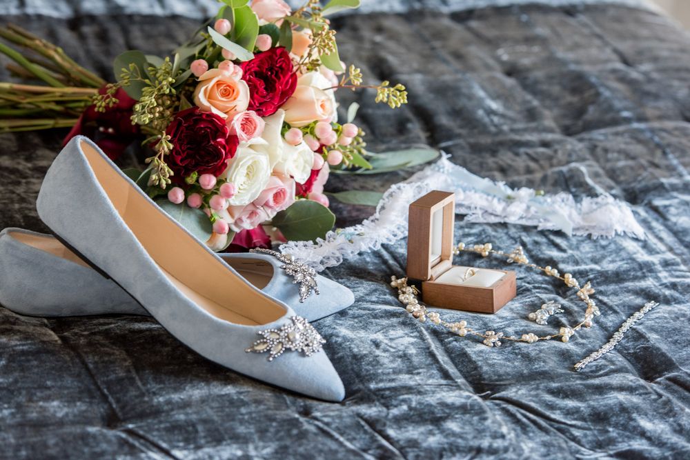Light blue bridal heels with crystal embellishment beside colorful bouquet, ring box, and pearl jewelry on gray velvet.