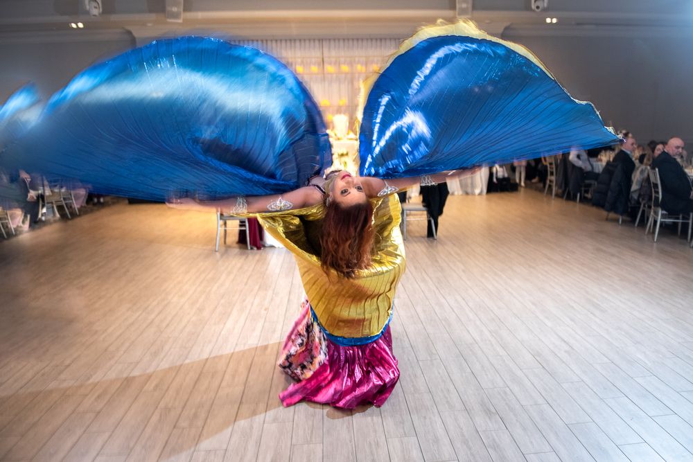 Belly dancer dramatically posing with large blue wings on elegant wedding reception dance floor.