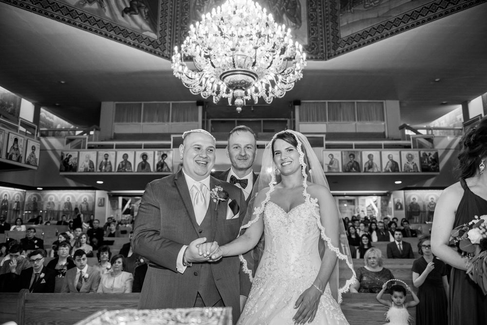 Newlyweds pose with officiant under grand chandelier in ornate Orthodox church after ceremony.