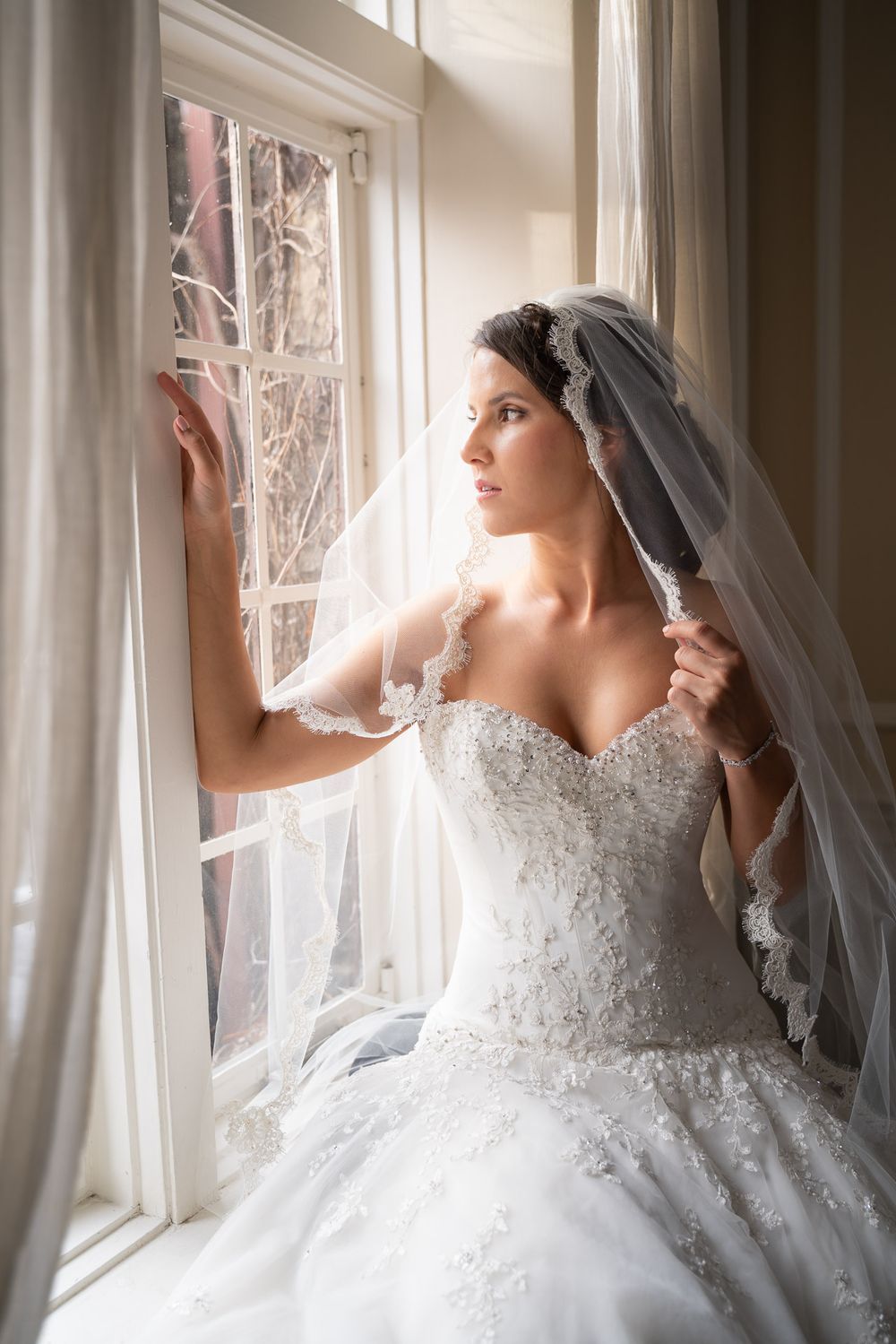 Beautiful bride in strapless lace gown lifts veil while gazing out sunlit window with sheer curtains.