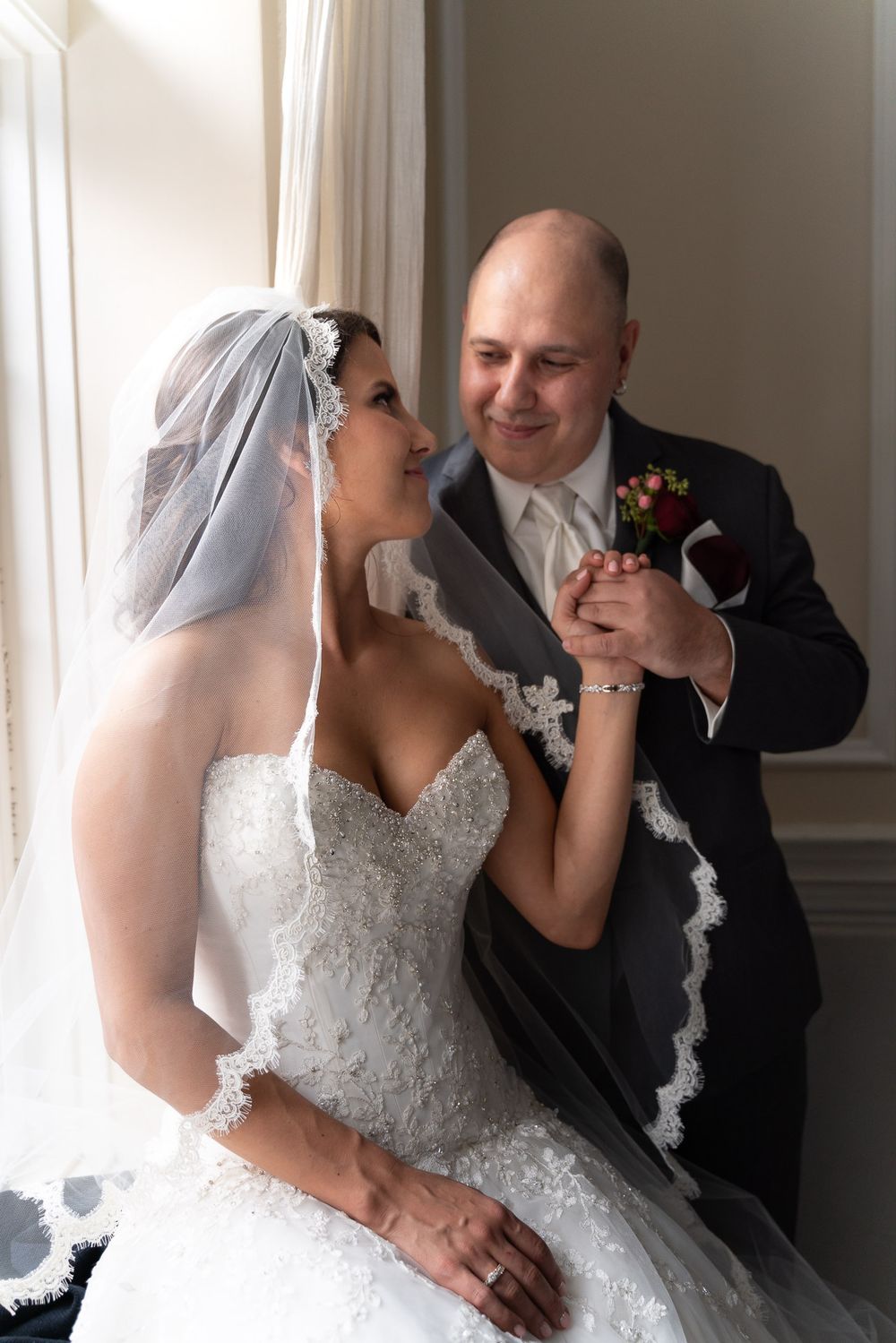 Bride and groom share a tender moment holding hands near a bright window on their wedding day.