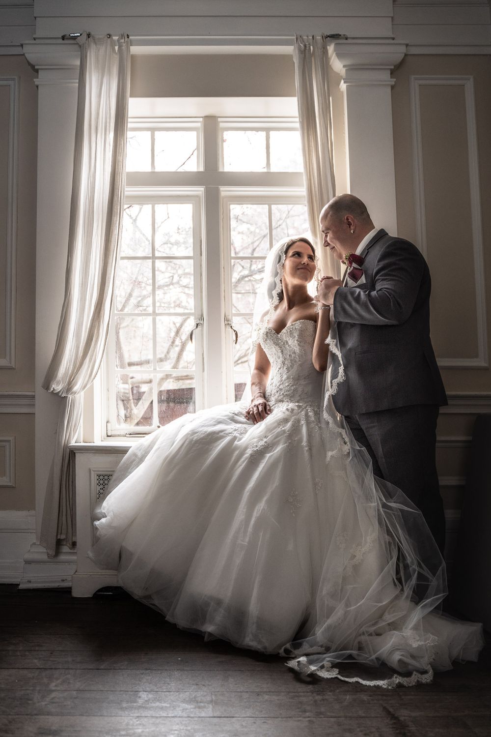 Bride in a ball gown sits on a window ledge as groom kisses her hand in an elegant room.
