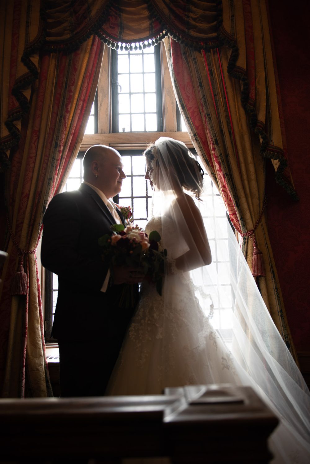 Silhouette of bride and groom holding bouquet before a grand window with gold and red drapes.