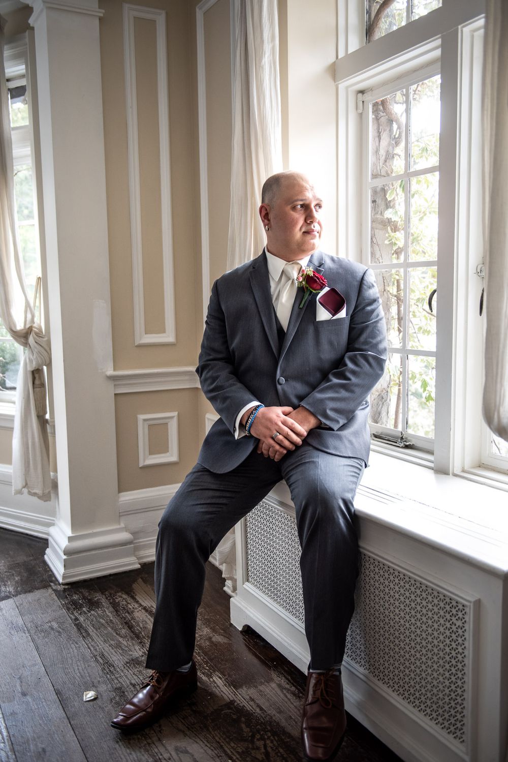 Dapper bald groom in gray three-piece suit with boutonniere sitting by large window in elegant venue.