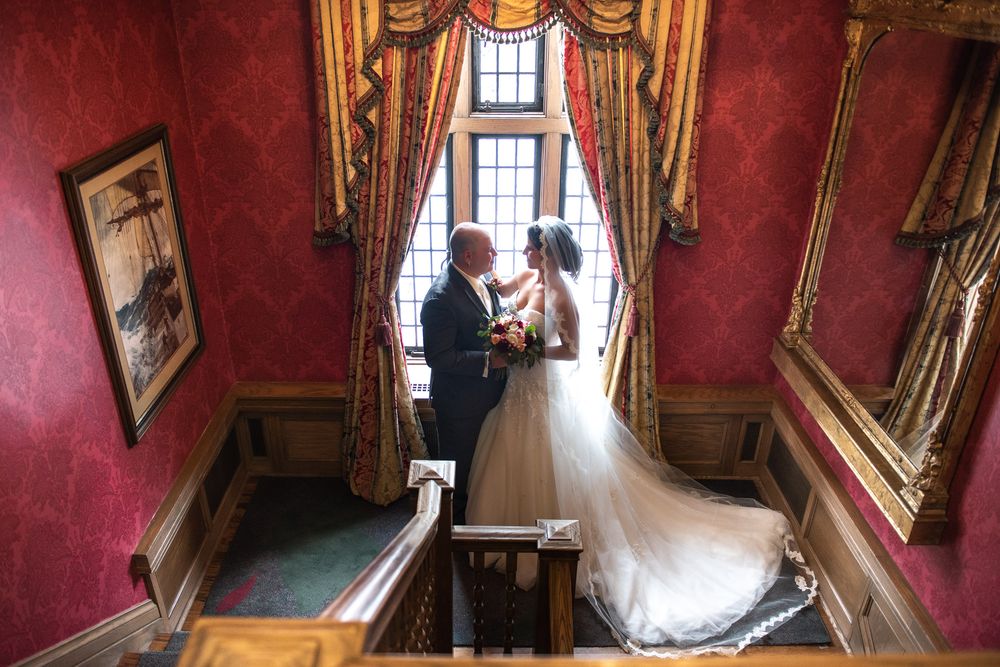 Bride and groom share intimate moment on grand staircase in opulent red-walled venue.