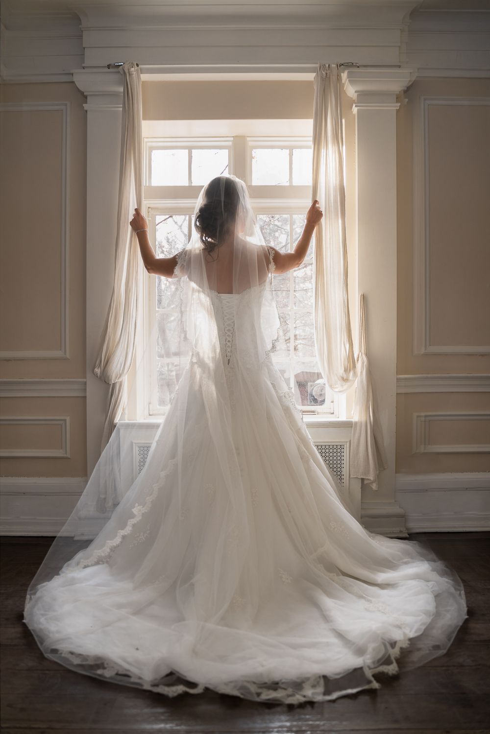 Elegant bride in white lace wedding gown with long train standing before sunlit window from behind.