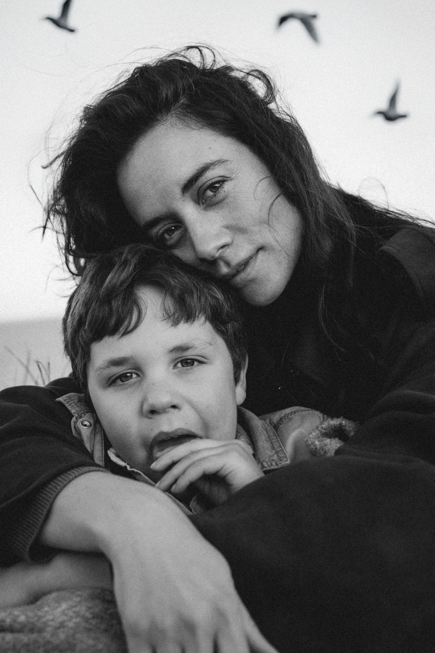 Black and white portrait of a mother and young boy resting together outdoors with birds flying in the background. Javea photographer
