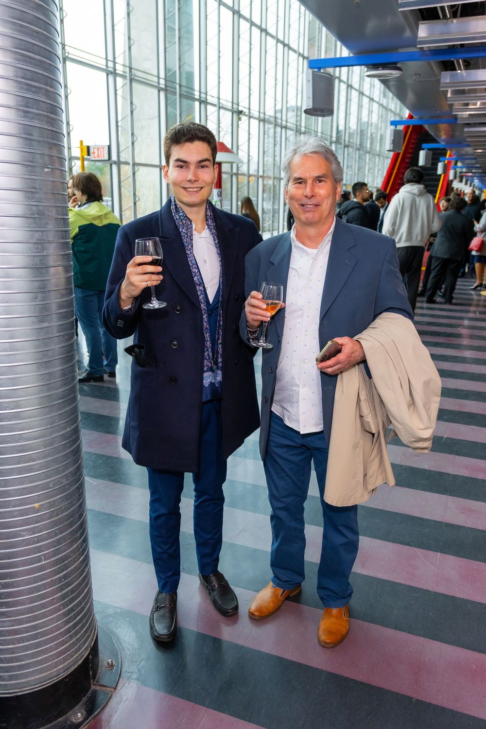 Two men smiling and holding wine glasses at an elegant indoor event with large windows.