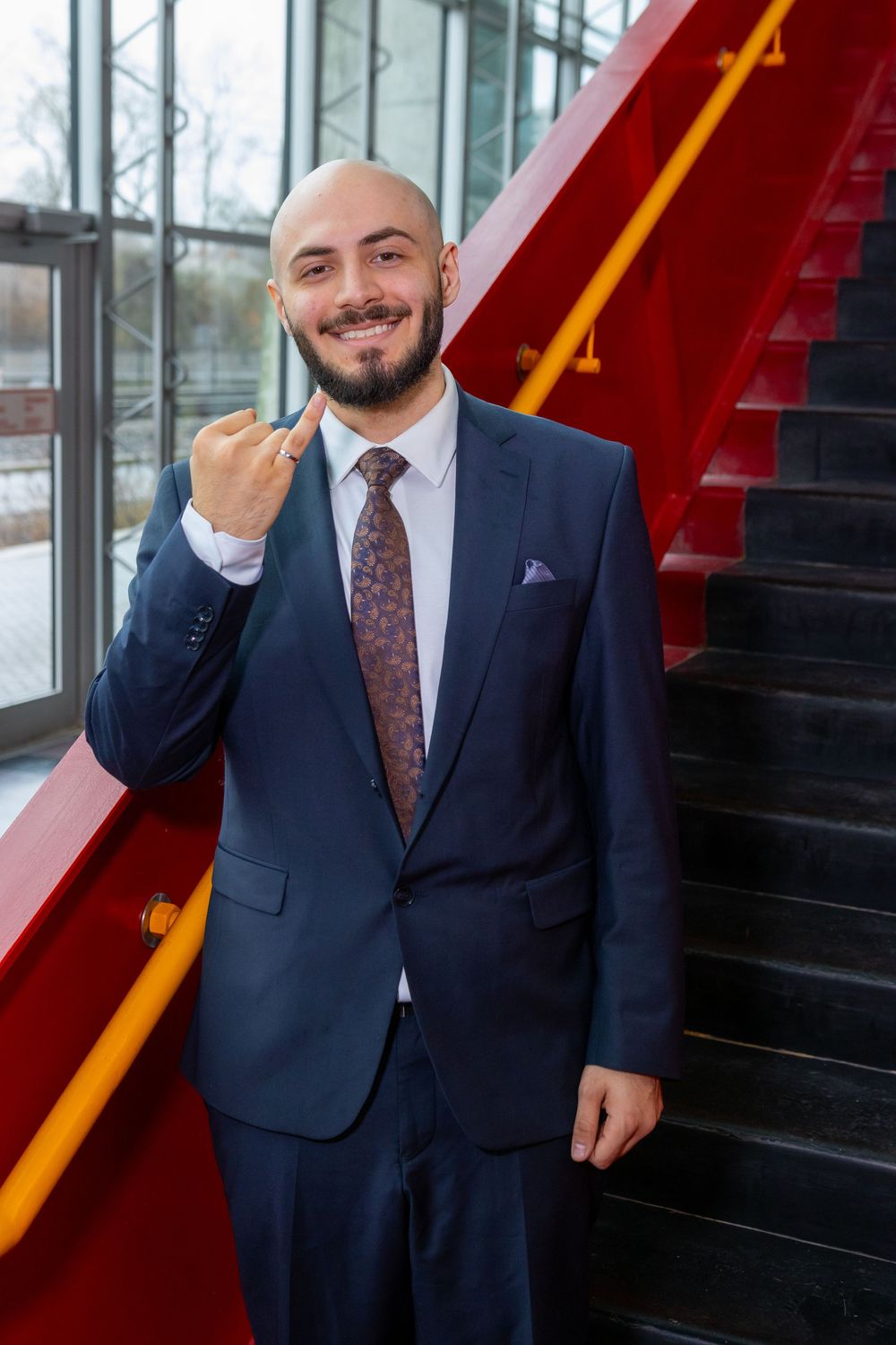 Smiling bald businessman in navy suit gesturing with finger near red staircase in modern office building.
