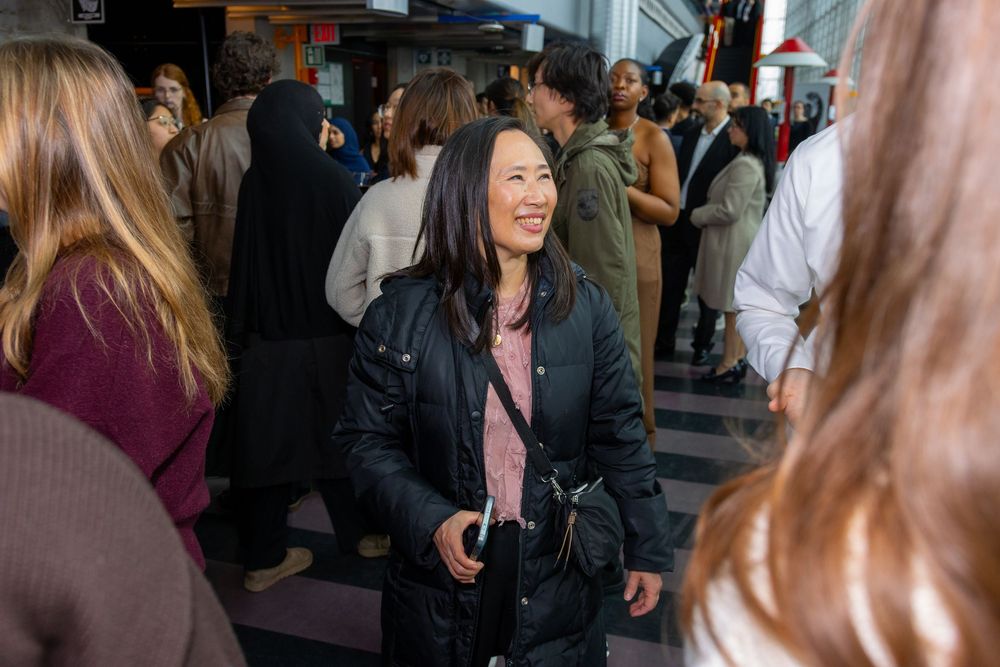 A smiling Asian woman in a black jacket mingles at a crowded indoor social event.