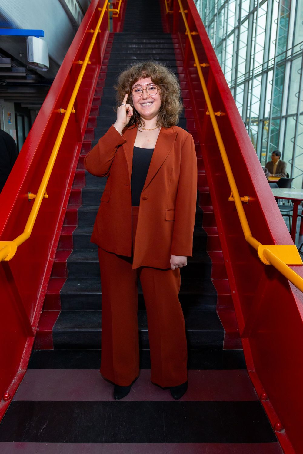 Woman in rust-colored pantsuit smiling on red staircase with yellow railings, talking on phone.