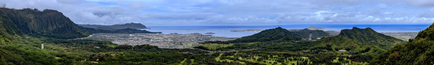 Panoramic view of Kaneohe Bay and lush green mountains on Oahu, Hawaii, with city below under partly cloudy skies.