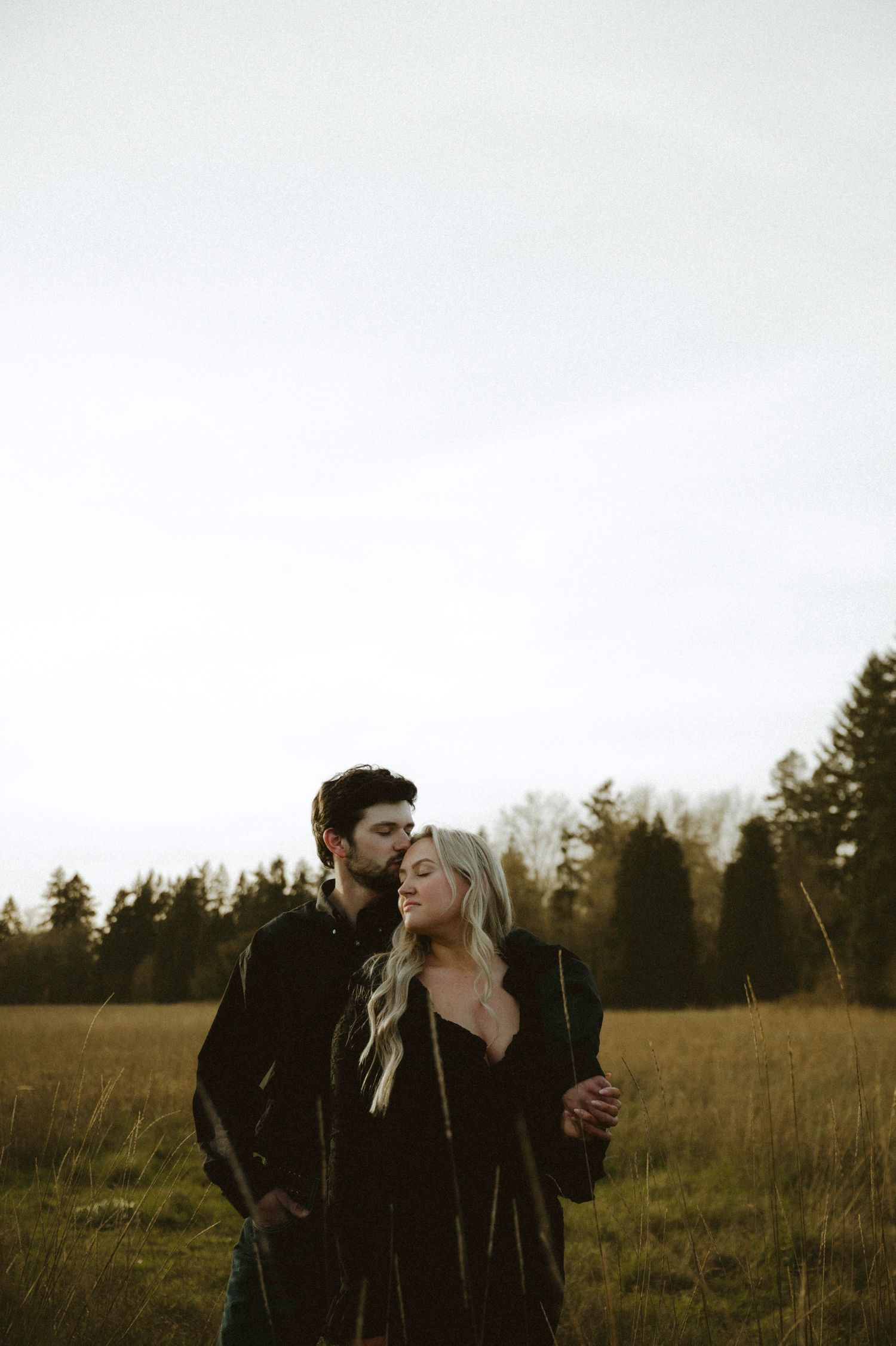 Couple embracing in a golden field at dusk, surrounded by tall trees, wearing dark clothing in a romantic outdoor setting.